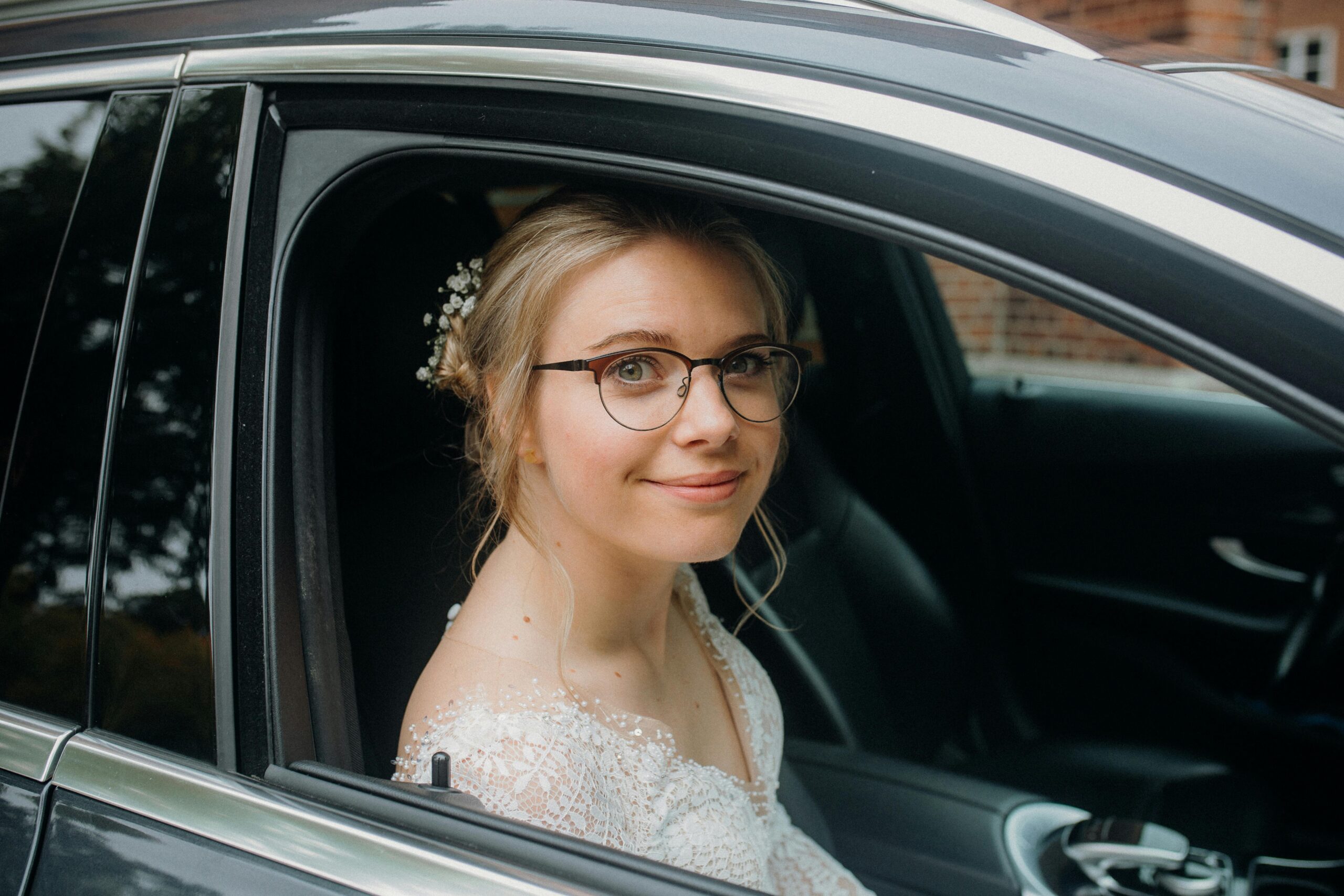 A joyful bride with blonde hair and eyeglasses smiles from a car window, capturing a gentle moment.