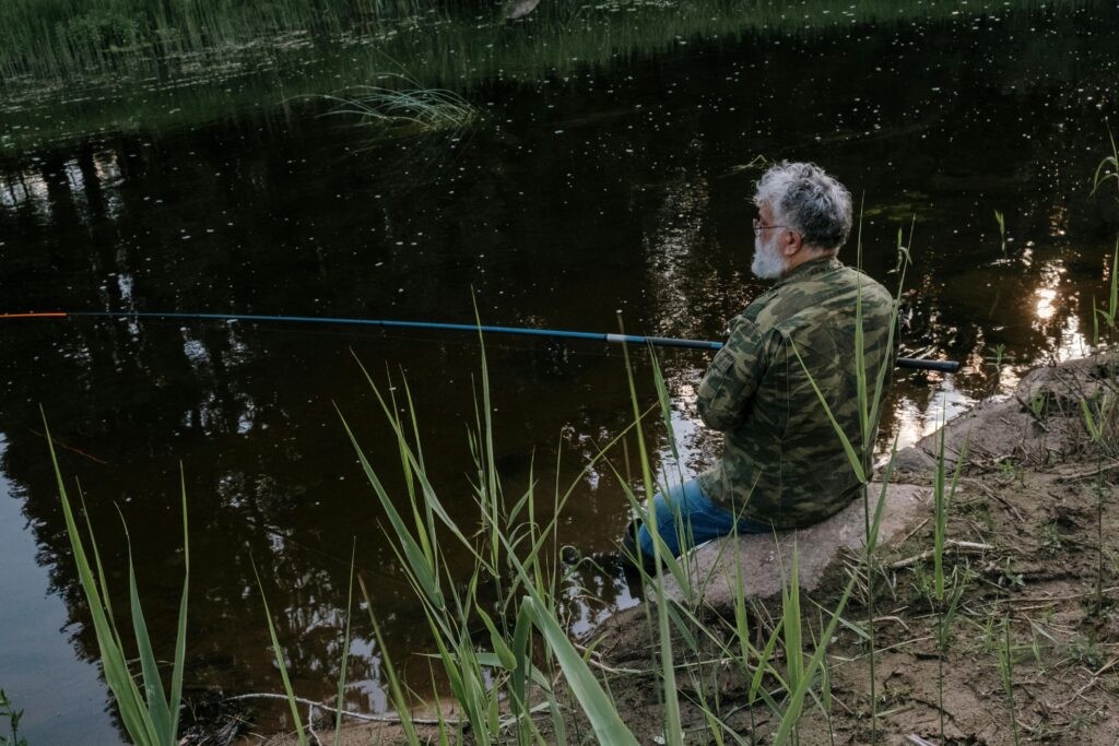 pexels-photo-4821080-4821080 Senior man in camo jacket enjoys quiet fishing at a serene riverbank surrounded by nature.
